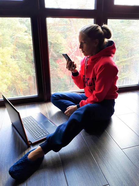 jovem menina de pele clara em casa trabalhando em um laptop e falando no telefone perto de uma grande janela
 - Foto, Imagem