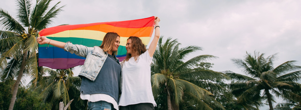 Δύο γυναίκες με σημαία ουράνιο τόξο στην παραλία σε φόντο φοινίκων - Φωτογραφία, εικόνα