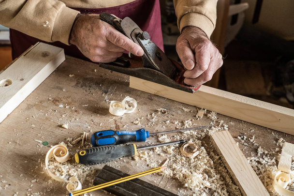 Carpenter man scraping curled wood scraps with hand plane tool and wooden plank. - Photo, Image