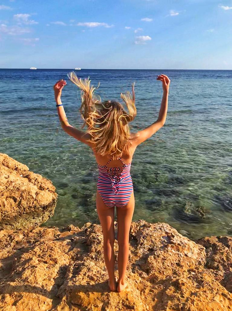 Felice bionda giovane ragazza con i capelli svolazzanti in un costume da bagno a righe in piedi sulla costa della spiaggia di corallo con le mani in alto e guardando il Mar Rosso in Egitto
 - Foto, immagini