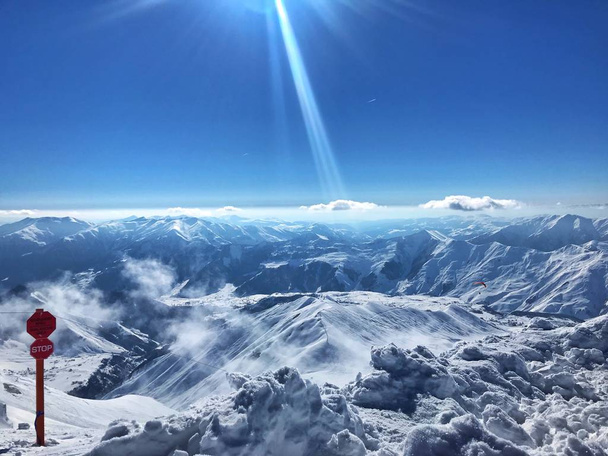 Vista para a montanha na Geórgia, nas montanhas do Cáucaso. Acima das nuvens, a vista das alturas das montanhas, perto de um punhado de neve e névoa clara, enquanto o sol brilha no céu azul. Sinal de parada na esquerda avisa de um penhasco
 - Foto, Imagem