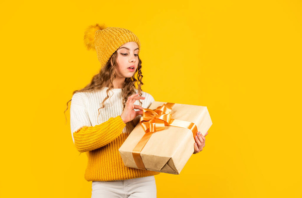 Christmas party time. cheerful little girl knitted hat and sweater. kid hold present box yellow background. xmas gift shopping sale. may your christmas be bright. happy winter holidays - Foto, Imagen