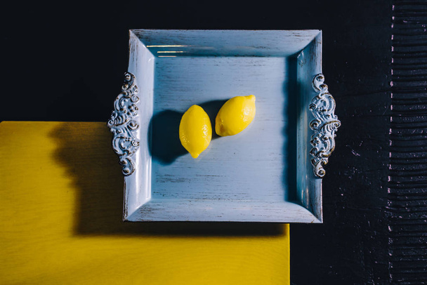 Μινιμαλισμός και λεμόνια στο τετράγωνο μπλε δίσκο σε κίτρινο και μαύρο φόντο. Κάτοψη με κενό για κείμενο. - Φωτογραφία, εικόνα