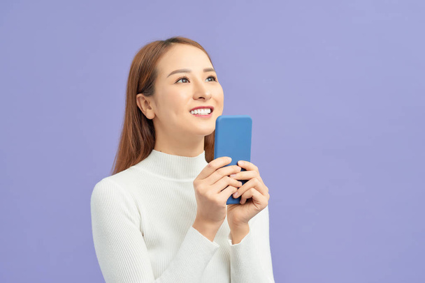 Sonriendo mujer asiática sosteniendo teléfono inteligente y mirando hacia otro lado sobre fondo violeta
 - Foto, Imagen
