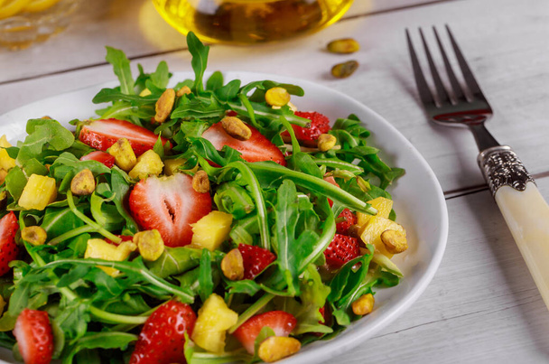 Healthy fresh salad with arugula, strawberries, pineapple and pistachios served on white plate on rustic wooden table. - Photo, Image