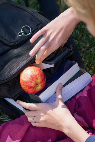 Student life concept. Black backpack stuffed with books. Hands holding books. Fresh apple for a healthy snack - Photo, Image