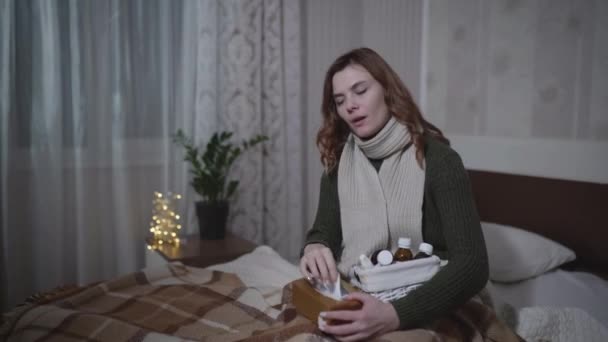 ill girl in warm clothes with stuffy nose sneezes and coughs while sitting on bed with napkins and medicines, sick female uses a drops from illness - Séquence, vidéo