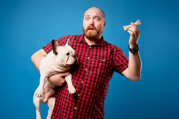 Surprised man in shirt holds bulldog and bone on empty blue background - Photo, Image
