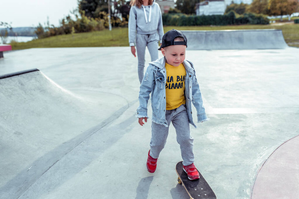 Cheerful happy little boy 3-5 years old, in summer on sports field city, learning to ride skateboard, emotions of joy, fun, relaxation and enjoyment. Casual wear, denim pants, baseball cap. - 写真・画像