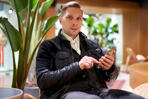 Male in leather jacket with smartphone looking at camera while sitting on chair in store against green plant - Фото, изображение