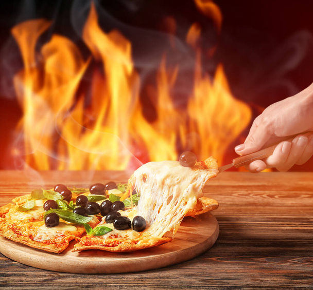 Жінка бере шматочок гарячої піци з оливками зі столу проти палаючого полум'я
 - Фото, зображення