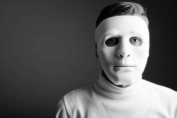 Studioaufnahme eines jungen Mannes mit weißem Rollkragenpullover und weißer Maske vor grauem Hintergrund in schwarz-weiß - Foto, Bild