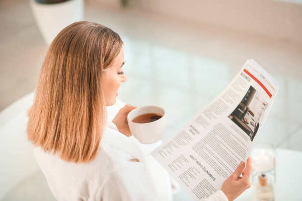 Beautiful young woman drinking tea and reading newspaper in bathroom - Photo, image