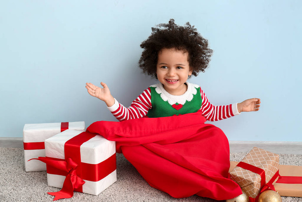 Маленька дівчинка в костюмі ельфа, з мішком Санта-Клауса і різдвяними подарунками сидять біля кольорової стіни. - Фото, зображення