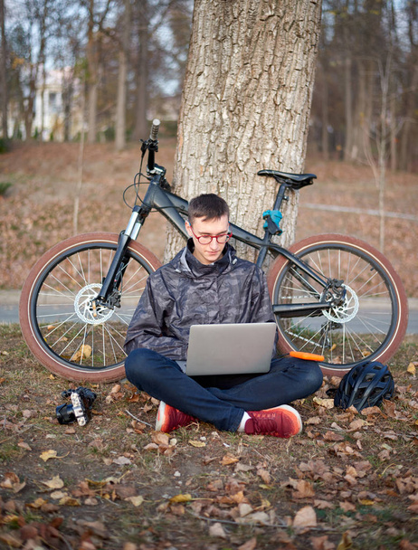 Young cyclist works at the computer in the park with a bicycle in the background - Fotó, kép