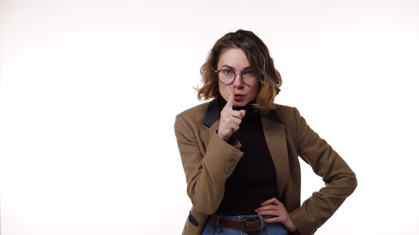 Angry, irritated emotions. Hush and keep silence. Caucasian brunette woman in glasses with finger on lips showing silent or hush gesture isolated on white background. Girl wearing black blazer and - Footage, Video