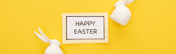 top view of greeting card with happy Easter lettering near Easter white bunnies on yellow colorful background, panoramic shot - Foto, afbeelding