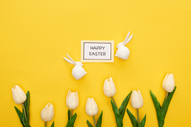top view of tulips, greeting card with happy Easter lettering near Easter white bunnies on yellow colorful background - Fotoğraf, Görsel