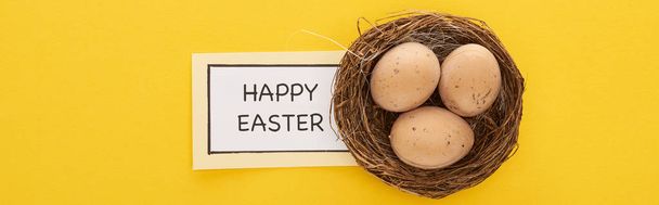 top view of greeting card with happy Easter lettering near chicken eggs in nest on yellow colorful background, panoramic shot - Photo, Image
