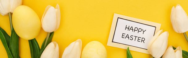 チューリップのトップビューとカラフルな黄色の背景に幸せなイースターの文字でカードの近くにイースターの卵を描いた、パノラマショット - 写真・画像