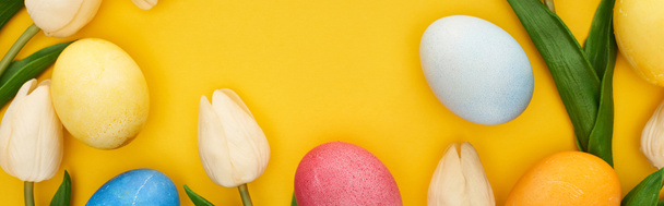 horní pohled na tulipány a malované velikonoční vajíčka na barevném žlutém pozadí s kopírovacím prostorem, panoramatický záběr - Fotografie, Obrázek