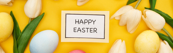 top view of tulips and painted Easter eggs around card with happy Easter lettering on colorful yellow background, panoramic shot - Fotografie, Obrázek