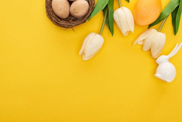 top view of tulips and chicken eggs in nest near Easter bunny on colorful yellow background - Photo, Image