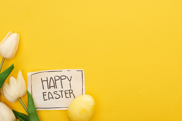 top view of Easter egg, tulips and greeting card with happy Easter lettering on yellow colorful background - 写真・画像