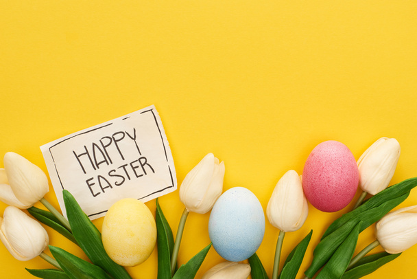 top view of Easter eggs, tulips and greeting card with happy Easter lettering on yellow colorful background - Photo, Image
