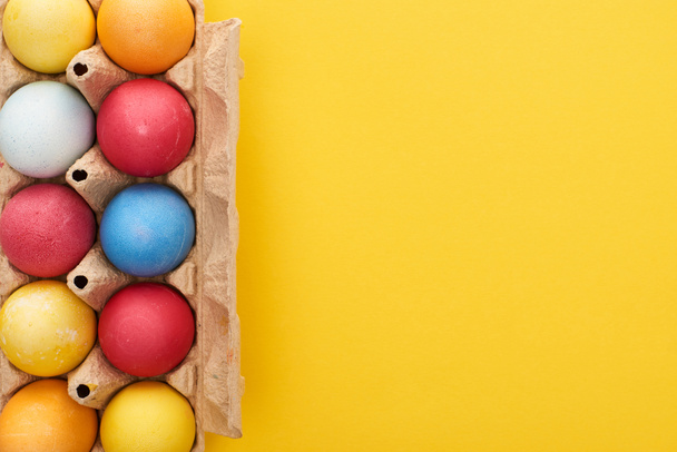 top view of multicolored painted Easter eggs in cardboard container on yellow background with copy space - Fotografie, Obrázek