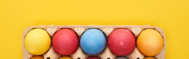 top view of multicolored painted Easter eggs in cardboard container on yellow background, panoramic shot - Foto, Imagem