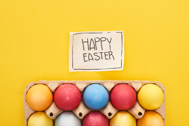top view of painted eggs in box and greeting card with happy Easter lettering on yellow colorful background - Foto, afbeelding