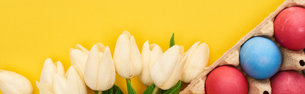 top view of tulips and multicolored painted Easter eggs in cardboard container on colorful yellow background, panoramic shot - 写真・画像