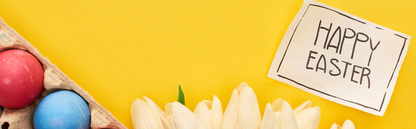 top view of painted eggs, tulips and greeting card with happy Easter lettering on yellow colorful background, panoramic shot - Fotó, kép
