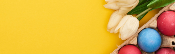 вид зверху на тюльпани і різнокольорові розмальовані великодні яйця в картонній тарілці на барвистому жовтому тлі, панорамний знімок
 - Фото, зображення