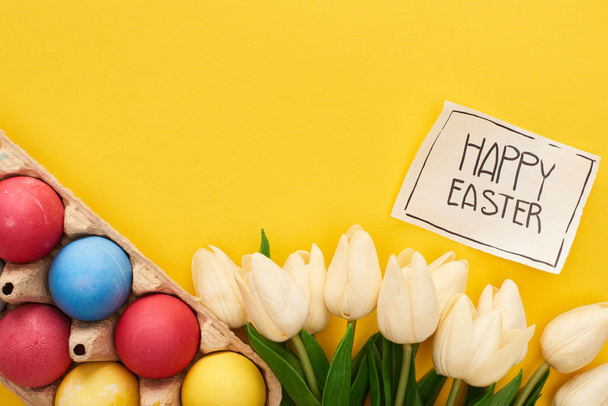 top view of painted eggs, tulips and greeting card with happy Easter lettering on yellow colorful background - Foto, afbeelding
