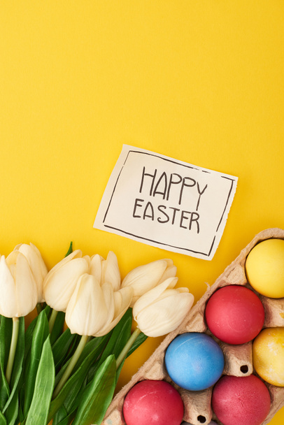 top view of painted eggs, tulips and greeting card with happy Easter lettering on yellow colorful background - Photo, Image