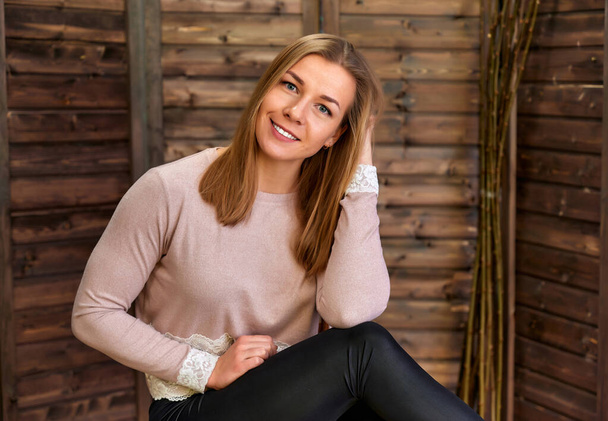 Loft style concept. Portrait of a pretty blonde girl in a pink sweater on a wooden background in the home interior. Cute looks and smiles at the camera, sitting on a chair straight - Foto, Bild