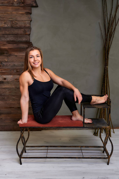 Portrait of a pretty fitness blonde girl in a tracksuit on an alternative background in vintage interior. Cute looks and smiles at the camera, sitting on a stand straight. Loft style concept. - Foto, Imagen