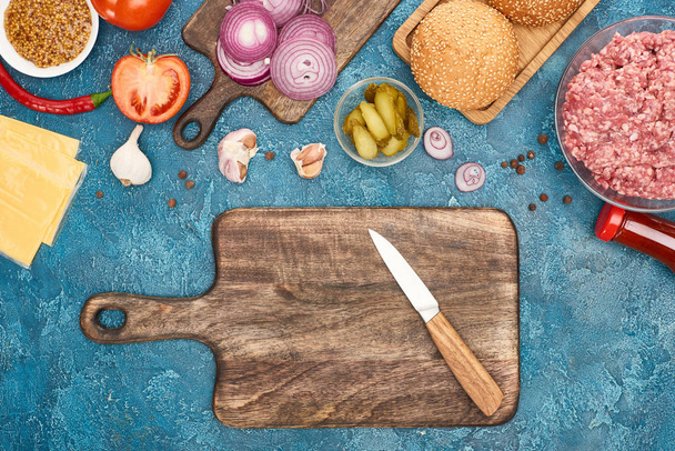 top view of fresh burger ingredients and wooden cutting board with knife on blue textured surface - Photo, Image