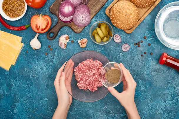 widok z góry kobieta dodając czarny pieprz do mielonego mięsa w pobliżu składników burger na niebieskiej powierzchni teksturowanej - Zdjęcie, obraz