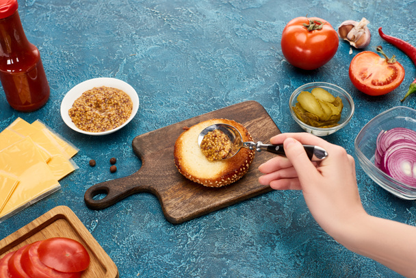 cropped view of woman adding mustard on bun on blue textured surface - Photo, Image