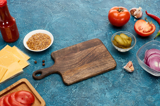 ingredientes frescos de la hamburguesa alrededor de tabla de cortar de madera en la superficie texturizada azul
 - Foto, imagen
