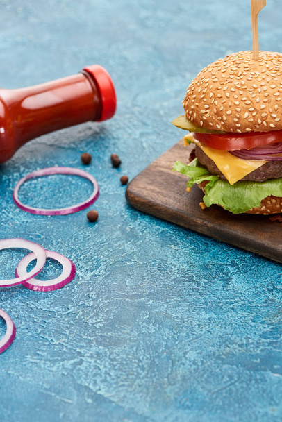 deliciosa hamburguesa con queso sobre tabla de madera cerca de cebolla y ketchup sobre superficie de textura azul
 - Foto, Imagen