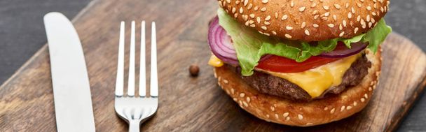 close up view of delicious fresh cheeseburger on wooden board with cutlery, panoramic shot - Foto, imagen