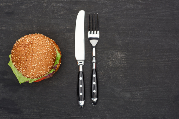 top view of delicious fresh burger on black wooden table with knife and fork - Photo, Image