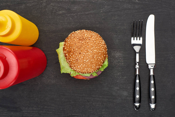 vista superior de delicioso hambúrguer fresco perto de talheres, ketchup e mostarda na mesa de madeira preta
 - Foto, Imagem