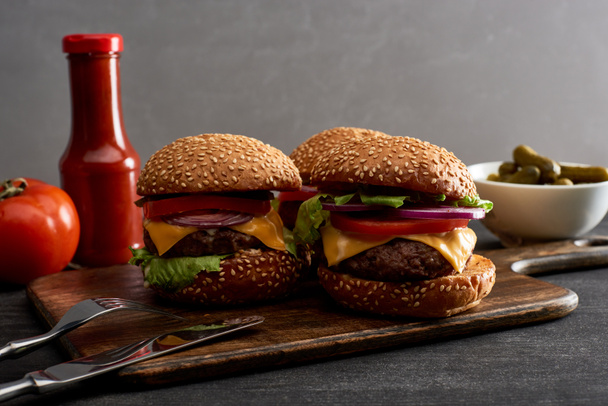 вкусные свежие мясные чизбургеры на деревянной доске возле столовых приборов, ингредиентов и кетчупа
 - Фото, изображение