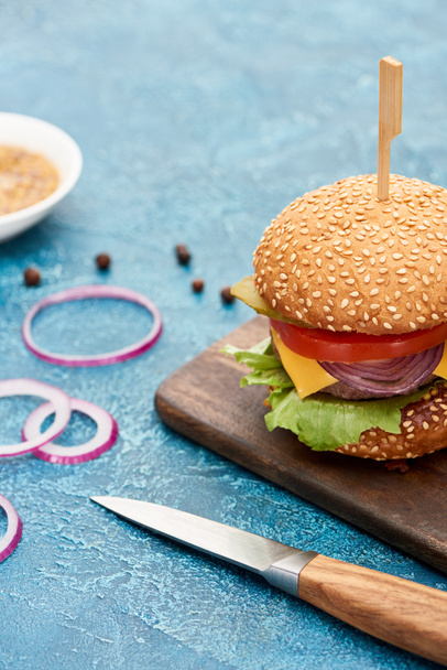 selective focus of knife and delicious cheeseburger on wooden board on blue textured surface - Foto, Bild