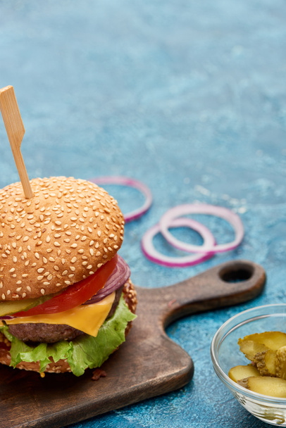 selective focus of delicious cheeseburger on wooden board near pickles on blue textured surface - Photo, Image